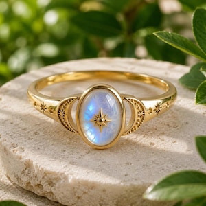 May include: A gold ring featuring an oval-shaped iridescent moonstone cabochon at the centre. The stone is accented with a small gold starburst emblem. The band displays crescent moon motifs and engraved star patterns along the sides.
