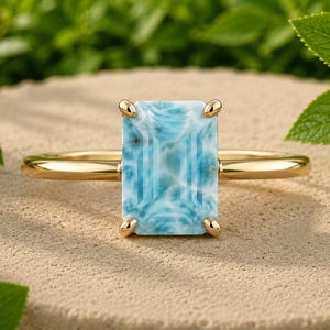 May include: A gold ring featuring a rectangular, emerald-cut larimar gemstone with a marbled light blue and white pattern. The stone is held in place by four gold prongs on a smooth, polished gold band, creating a clean and simple jewelry piece.