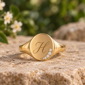 Peut inclure: Une bague chevalière en or avec un plateau circulaire poli gravé d'une lettre H en cursive. Un accent en forme de croissant serti de cinq petites pierres rondes et transparentes est placé sur le bord inférieur droit du plateau.