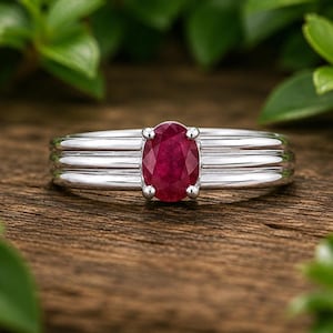 May include: A silver-toned ring featuring a central oval-cut deep red ruby gemstone held by four prongs. The band is constructed with three distinct horizontal parallel ridges that wrap around the finger, creating a layered metal design.