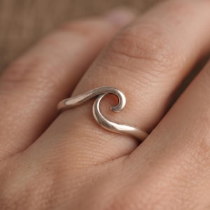 Può includere: Un anello in argento lucido caratterizzato da un design a onda continuo e liscio che si curva a spirale al centro. La fascia è sottile e segue il contorno naturale del dito, mostrando una finitura metallica e riflettente su tutto il pezzo.