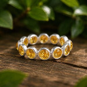 May include: A silver eternity band featuring a continuous row of round, faceted yellow citrine gemstones set in individual metal bezels. The ring displays a uniform design with bright, warm-toned stones encircling the entire circumference of the band.