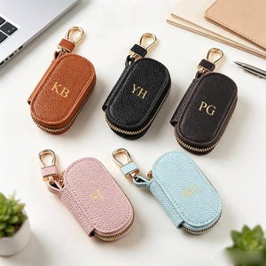 May include: Five oval-shaped key cases with textured surfaces and gold-tone zippers. Each case features a gold-tone metal clasp and is personalized with two gold-lettered initials: KB in tan, YH in black, PG in dark brown, SF in pink, and SG in light blue.