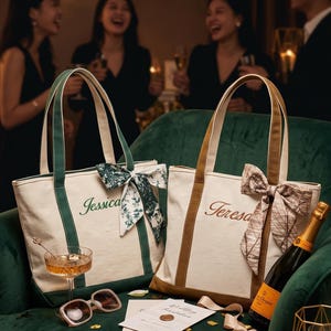 Può includere: Due borse tote in tela con doppi manici e fiocchi in tessuto decorativi. Una borsa presenta un bordo verde e il nome Jessica ricamato in verde. L'altra borsa ha un bordo marrone e il nome Teresa ricamato in marrone.