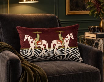 May include: A rectangular velvet throw pillow in deep burgundy featuring two riders in wide-brimmed hats on spotted white horses. The lower section displays a black and white zebra stripe pattern, separated from the burgundy by a thin green line.