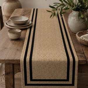 May include: A rectangular table runner featuring a textured, natural tan woven base with two distinct, parallel black border stripes running along the length of the fabric, creating a framed effect on the surface of the wooden dining table.