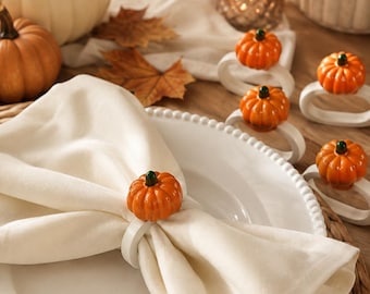 May include: A set of white napkin rings, each featuring a glossy orange pumpkin with a small green stem. One ring secures a folded white cloth napkin on a white beaded plate, while several other identical rings are arranged nearby on the table.