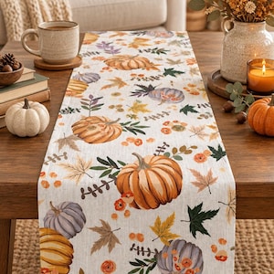 Puede incluir: Un camino de mesa rectangular con un patrón repetitivo de calabazas naranjas y moradas, hojas de otoño en tonos amarillos, marrones y verdes, y pequeñas bayas naranjas impresas sobre una tela de color beige claro con textura.