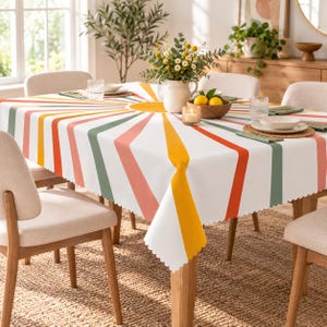 May include: A rectangular tablecloth featuring a white background with a radiating sunburst pattern in shades of orange, yellow, and sage green. The fabric has a scalloped edge and is draped over a dining table set with plates, glasses, and a vase.