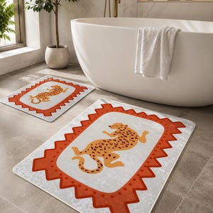 May include: Two rectangular bath mats featuring a central light gray field with a stylized orange leopard illustration. Each mat is framed by a wide, jagged-edged orange border with a repeating dot pattern along the inner edge of the border.