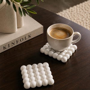 Pode incluir: Dois porta-copos quadrados brancos com uma textura de bolhas em relevo feita de pequenas esferas dispostas em grade. Um porta-copos sustenta uma caneca de cerâmica com café preto, enquanto o outro está vazio sobre uma superfície de madeira.