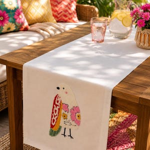 Pode incluir: Um caminho de mesa branco com um design de pássaro bordado. O pássaro tem um corpo cor de creme, uma asa vermelha com padrões brancos, uma cauda verde e detalhes florais rosa e amarelos na lateral. O caminho está sobre uma mesa de madeira.