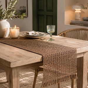 May include: A rectangular brown table runner featuring an open-weave, textured pattern with fringe detailing on the ends. The runner is crafted from a natural-toned fiber material and is draped across the center of a wooden dining table.