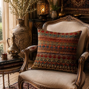 Può includere: Un cuscino decorativo con un motivo geometrico nei toni del rosso, arancione, blu e marrone poggia su una sedia imbottita color crema con dettagli in legno ornati. La scena evoca un'estetica bohémien e accogliente.
