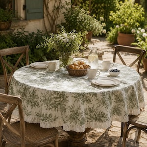 Puede incluir: Una mesa redonda cubierta con un mantel blanco con un patrón floral verde y un borde de encaje blanco. La mesa está puesta con platos blancos, tazas, un tazón pequeño de arándanos, una cesta de mimbre con pan y un florero de cristal.