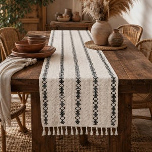 Pode incluir: Um caminho de mesa na cor creme com padrões geométricos pretos verticais e franjas em ambas as extremidades. O caminho está estendido sobre uma mesa de madeira, exibindo uma construção de tecido texturizado com desenhos lineares repetidos.