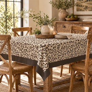 Könnte beinhalten: Eine rechteckige Tischdecke mit einem beigen und dunkelbraunen Leopardenmuster und einem einfarbigen dunkelbraunen Rand an der Unterkante. Der Stoff liegt auf einem Holztisch und bedeckt die Oberseite sowie die Seiten.