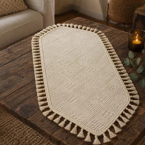 May include: An oval-shaped cream-colored table runner featuring a textured geometric knit pattern and a decorative border of small, uniform tassels along the entire edge. The runner is crafted from a thick, woven fabric with a consistent light tone.