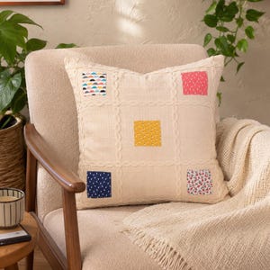 May include: A square cream-colored throw pillow featuring a grid of raised cable-knit lines. Five small, colorful fabric patches with distinct patterns are sewn onto the pillow surface in a scattered arrangement, including yellow, red, and blue.
