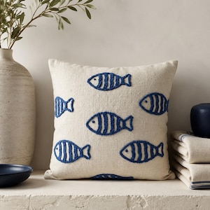 May include: A square throw cushion with a natural beige linen-effect fabric base featuring a repeating pattern of embroidered dark blue fish. Each fish has a simple striped design created with a raised, textured punch needle or tufted stitch technique.