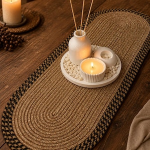 May include: An oval braided table runner featuring a tan center and a black and tan patterned border. A white circular tray holds a white ribbed diffuser with three reeds, a white ribbed candle, and a small white bowl filled with white stones.