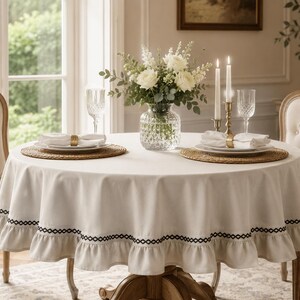 May include: A round off-white tablecloth with a ruffled edge featuring a black geometric embroidered trim. The tablecloth is draped over a circular table set with two woven placemats, white napkins, crystal glasses, and a central floral vase.