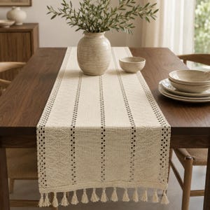 Pode incluir: Um caminho de mesa na cor creme com textura tecida, padrões de listras verticais, detalhes intrincados em forma de diamante e uma borda decorativa com franjas e pequenas borlas penduradas na extremidade do tecido sobre uma mesa de madeira.