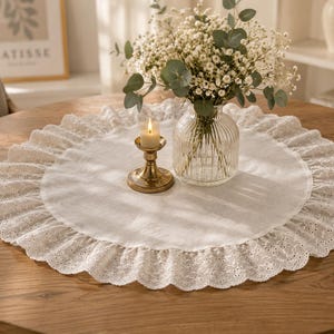 May include: A round white fabric table mat featuring a wide, ruffled border with intricate eyelet embroidery. The mat is placed on a wooden surface and holds a clear, ribbed glass vase filled with white flowers and a brass candle holder.