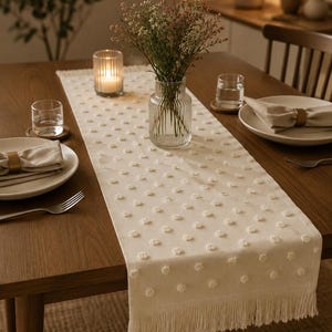 Pode incluir: Um caminho de mesa na cor creme com um padrão repetitivo de pequenos pontos tufados em relevo, finalizado com franjas longas e delicadas em ambas as extremidades. O caminho é colocado sobre uma mesa de madeira, oferecendo uma superfície.