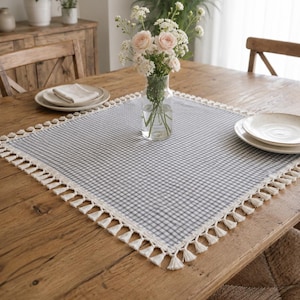 May include: A square table topper featuring a grey and white gingham check pattern with a decorative cream-colored fringe trim along all four edges. The fabric is laid flat on a wooden table surface, centered beneath a clear glass vase of flowers.
