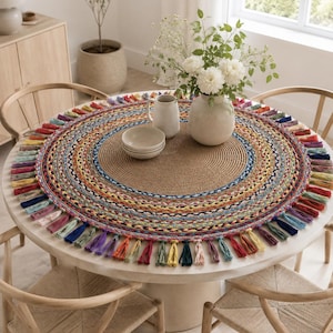 Pode incluir: Um tapete de mesa redondo com um centro castanho sólido feito de corda enrolada, rodeado por anéis concêntricos de tecido entrançado em várias cores, incluindo azul, amarelo, vermelho e verde, rematado com uma franja de borlas de tecido multicolor.