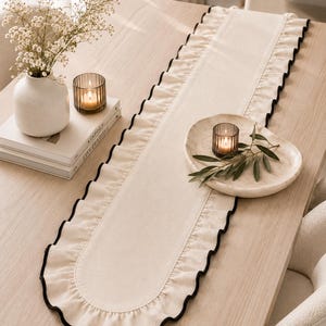 Puede incluir: Un camino de mesa largo de tela color crema con extremos redondeados y un borde con volantes fruncidos a lo largo de todo el perímetro. El volante está rematado con un fino detalle de borde negro en contraste que perfila la forma.