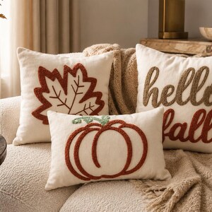 May include: Three cream-coloured throw pillows featuring textured embroidery. One square pillow displays a rust-coloured maple leaf, another square pillow features the text "hello fall" in brown and rust, and a rectangular pillow shows a pumpkin.