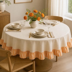 May include: A round cream-coloured tablecloth with a ruffled orange and white striped border. The table is set with white ceramic bowls, plates, clear glass tumblers, gold-toned cutlery, and a white vase filled with orange ranunculus flowers.