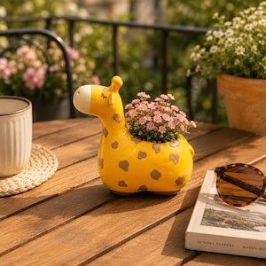 以下が含まれることがあります： ピンクの花で満たされた、茶色の斑点のある黄色のキリン型のプランター。セラミックプランターは木製のテーブルの上にあり、カップとサングラスが近くにあります。キリンは白い顔と小さな黒いディテールを持っています。