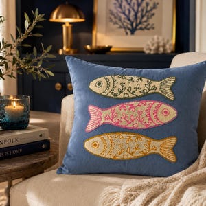 May include: A square blue throw pillow featuring three embroidered fish stacked vertically. The top fish is cream, the middle fish is pink, and the bottom fish is yellow. Each fish has a detailed floral pattern stitched into its body.