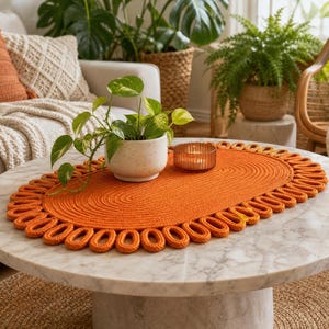 Peut inclure: Un set de table ovale orange avec un centre enroulé et une bordure décorative à boucles. Le set est posé sur une table ronde en marbre blanc, supportant un petit pot en céramique blanche mouchetée avec une plante verte et un bougeoir.