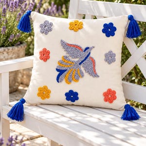 Puede incluir: Un cojín cuadrado de color blanco roto con un pájaro bordado en el centro en tonos gris, naranja y azul. El cojín está decorado con ocho pequeñas flores de ganchillo en amarillo, azul, gris y naranja, y tiene una borla azul en cada esquina.