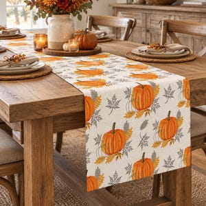 Peut inclure: Un chemin de table rectangulaire orné d'un motif répétitif de citrouilles orange, d'épis de blé jaunes et de feuilles d'érable grises sur un fond en tissu crème clair, disposé sur une table à manger en bois avec des couverts et un centre de table.