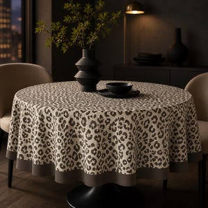 May include: A round tablecloth featuring a beige and dark grey leopard print pattern with a solid dark grey border along the bottom edge. The fabric drapes over a circular table, covering the surface completely with a consistent, repeating animal print.