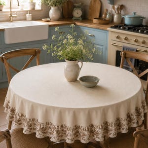 Op de afbeelding: Een rond, crèmekleurig tafelkleed met een geplooide rand met een bruin bloemenpatroon. De stof heeft een textuur en hangt over een ronde tafel, waarop een keramieken kan met witte bloemen en een klein kommetje zijn geplaatst.