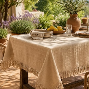 May include: A beige rectangular tablecloth with a subtle grid pattern and long fringe trim along the edges. The fabric has a textured weave, draping over the corners of a wooden table. The tablecloth is neutral in color and features a consistent square grid design throughout the entire surface area of the textile.