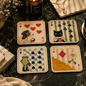 Puede incluir: Un juego de cuatro posavasos cuadrados con diseños de cartas de juego. Uno muestra un perro con cinco corazones, otro un gato con siete tréboles, un tercero una rana con ocho picas y el último un personaje de margarita con un símbolo de sota de diamantes.