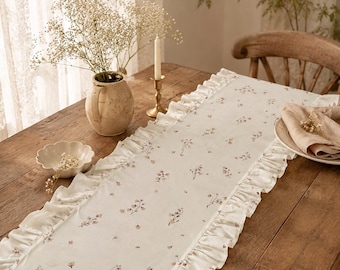 May include: A rectangular off-white table runner featuring a delicate floral pattern with small brown and pink blossoms. The runner is finished with gathered fabric ruffles along both long edges, creating a textured border along the length.