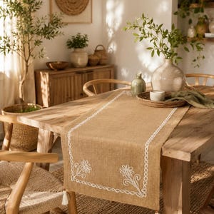 May include: A rectangular tan burlap table runner featuring a white embroidered floral pattern along the border and white tassels attached to the corners. The fabric has a visible woven texture and is draped across a wooden dining table.
