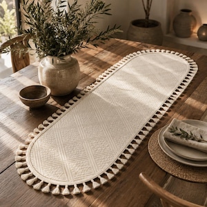 May include: An oval-shaped off-white table runner featuring a textured diamond pattern across its surface. The edges of the runner are finished with a decorative fringe of small, evenly spaced tassels that extend around the entire perimeter.
