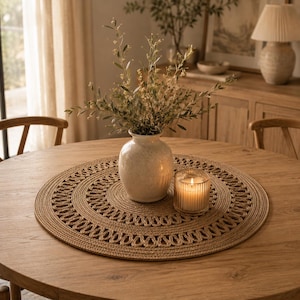 Peut inclure: Un set de table rond en jute tressé avec un motif de bordure tressée décoratif. Au centre du set se trouve un vase en céramique blanc cassé moucheté contenant des branches feuillues vertes, accompagné d'un petit bougeoir en verre strié.