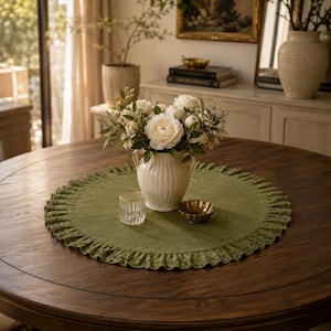 Op de afbeelding: Een ronde olijfgroene stoffen placemat met een geplooide rand en gedetailleerde kanten afwerking. In het midden staat een witte, gegroefde keramische vaas met witte bloemen en groen, vergezeld door een helder glazen beker en een kleine gouden kom.