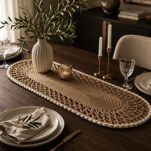 May include: An oval-shaped table runner featuring a central solid tan woven section surrounded by a decorative brown macrame lattice border and finished with a thick cream-colored braided edge, displayed on a dark wooden dining table.