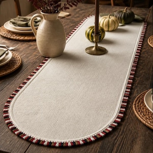 May include: An oval-shaped beige table runner featuring a textured fabric surface and a decorative border trim composed of alternating short vertical threads in black, white, red, and dark brown colours, creating a distinct multi-coloured edge.